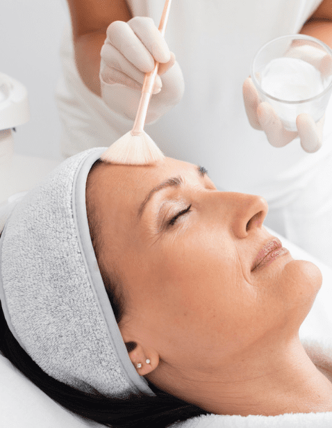 Professional aesthetician applying a facial peeling mask to a middle-aged woman who is relaxing with closed eyes in a salon.