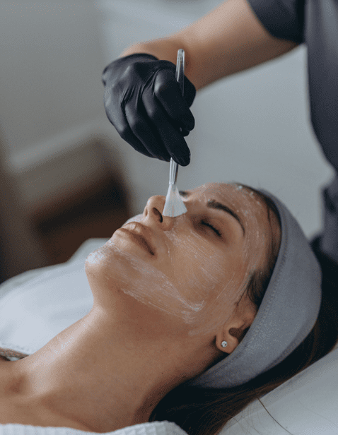 A professional wearing black gloves applies a facial mask to a relaxed woman lying on a spa bed with a gray headband.