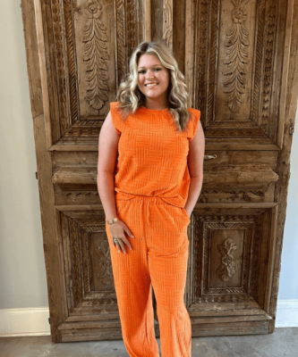 Smiling aesthetician in bright orange outfit standing in front of a carved wooden door at a spa, representing professional skincare services in Memphis.