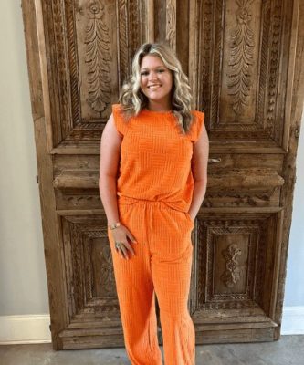 Smiling aesthetician in bright orange outfit standing in front of a carved wooden door at a spa, representing professional skincare services in Memphis.