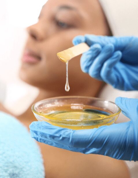 Aesthetician wearing blue gloves prepares warm wax for full face waxing treatment on a woman with dark skin, emphasizing skincare and hair removal services.
