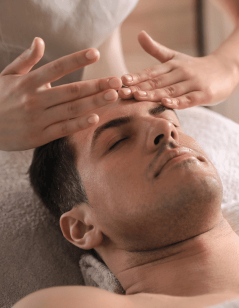 A man receiving a relaxing Gentlemen’s Facial, with a professional gently massaging his forehead while he lies with eyes closed.