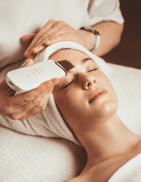 A woman receiving the IOIS Signature Facial with a professional using a skin exfoliation device on her forehead while she relaxes with eyes closed and a towel wrapped around her head.