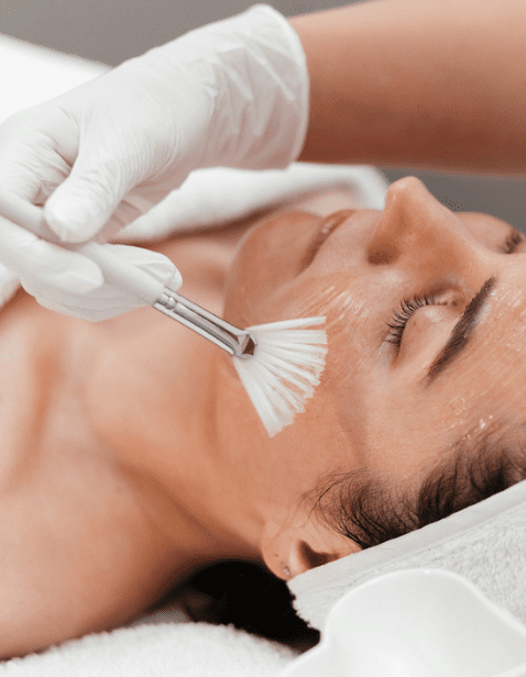 A professional wearing gloves applying a facial chemical peel with a brush to a relaxed woman lying down with eyes closed and a towel wrapped around her head.