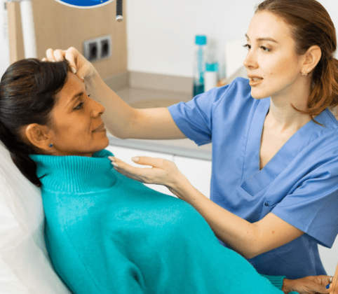A licensed skin care professional in blue scrubs conducting a facial consultation with a client wearing a teal sweater in a spa treatment room.