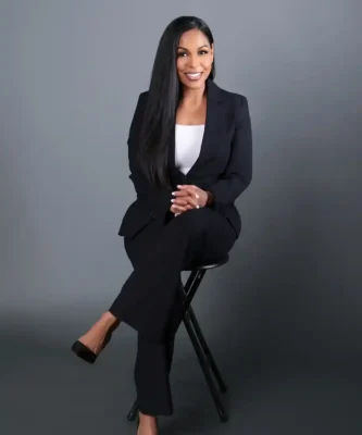 Professional portrait of Jonel Feliciano, founder of Isle of Indulgence Spa, wearing a black blazer and white top, smiling confidently with arms crossed against a blurred natural background.